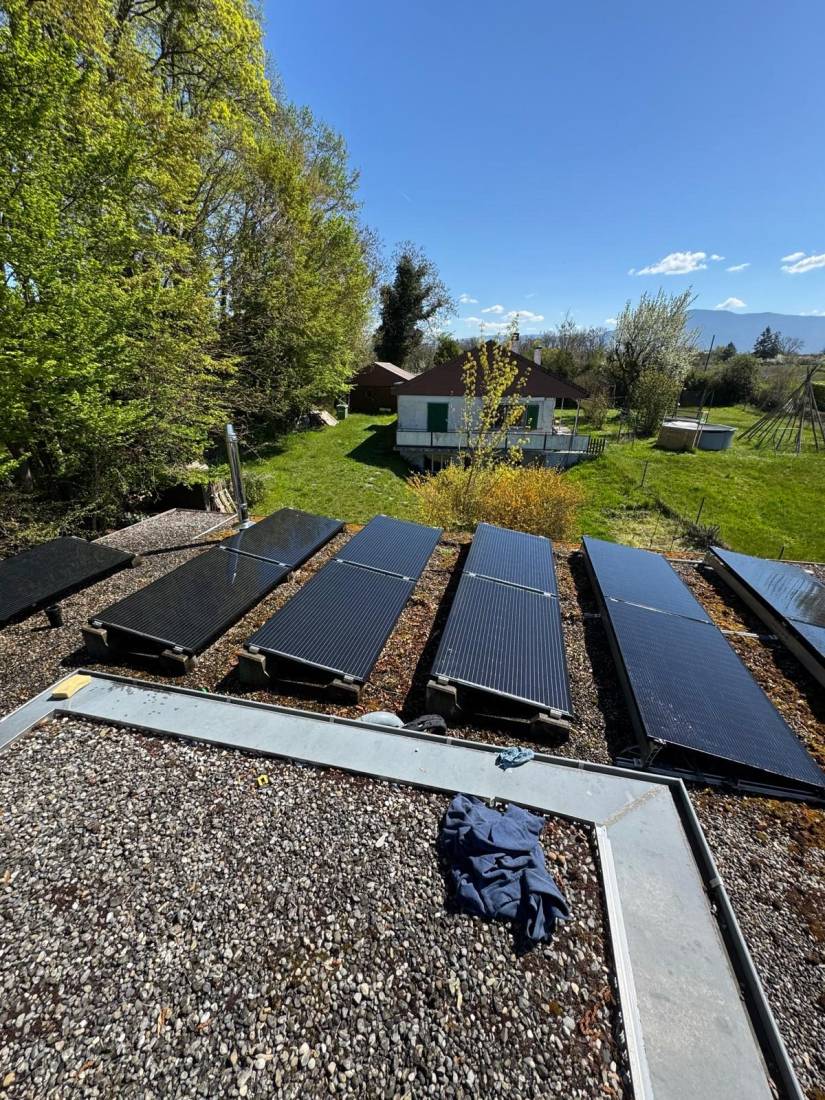 Panneaux solaires sur supports inclinés lestés au gravier sur un toit plat donnant sur un jardin en Suisse romande