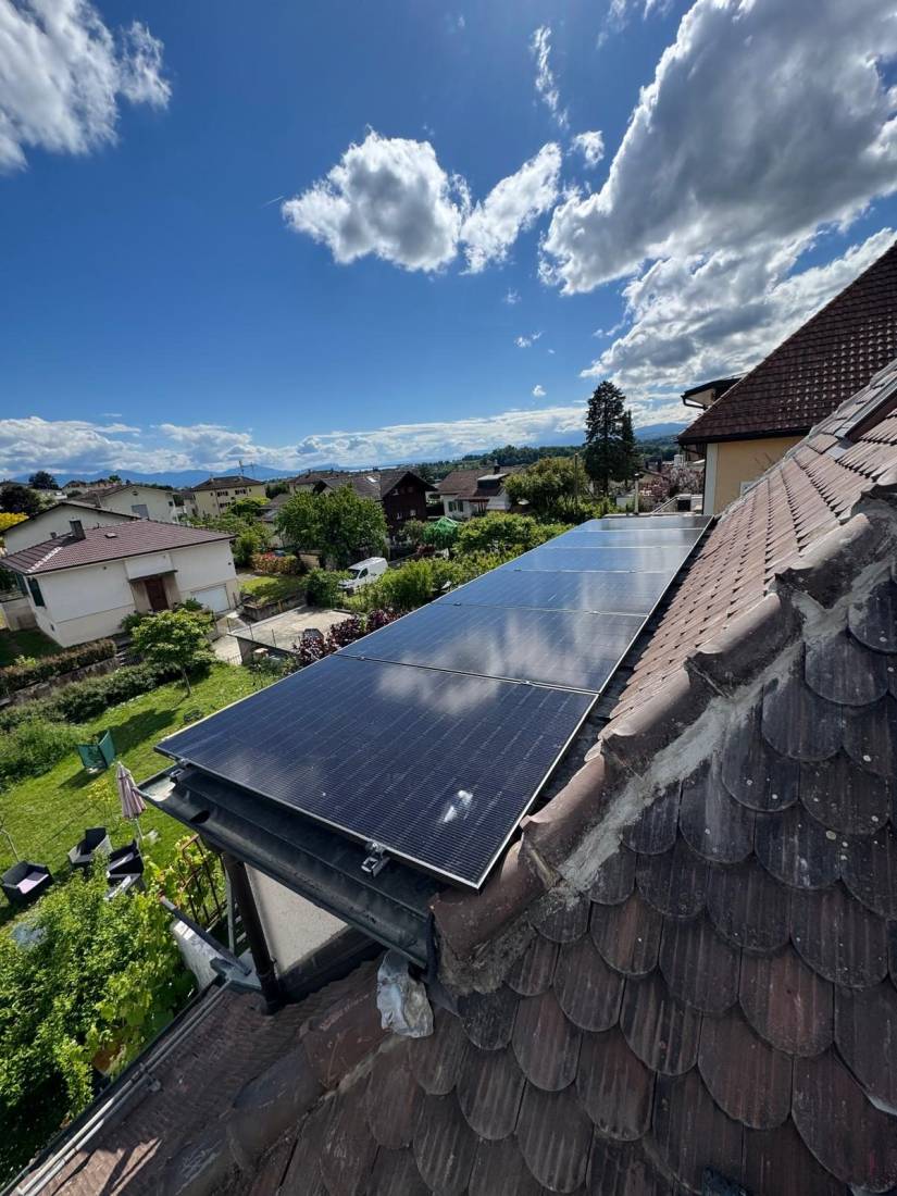 Rangée de panneaux solaires sur toiture en tuiles avec vue sur un village et ciel bleu en Suisse romande